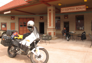 white motorcycle in foreground with rider sitting at an outdoor table under a coffee shop sign
